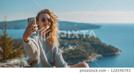 A beautiful woman, smiling, radiating happiness, takes selfie overlooking a stunning coastal landscape with blue waters, lush greenery, perfect for themes of travel, freedom, enjoying nature's beauty 128340763