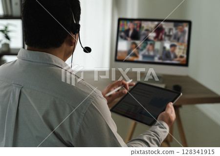 Virtual Meeting and virtual conference person participating virtual meeting using headset, tablet, and computer monitor displaying multiple people 128341915