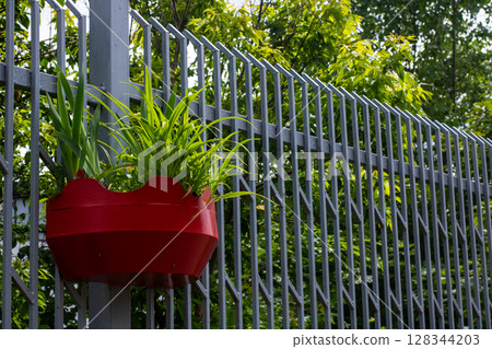 A red planter with various plants hangs from a sturdy metal fence 128344203