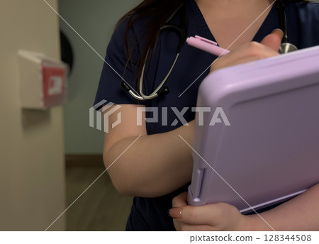 Nurse Writing on Clipboard in Hallway with Stethoscope Around Neck Nurse Writing on Clipboard in Hallway with Stethoscope Around Neck 128344508
