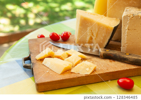 Chunky pieces of hard cheese with knife and cherry tomatoes on wooden board in bright outdoor setting 128346485