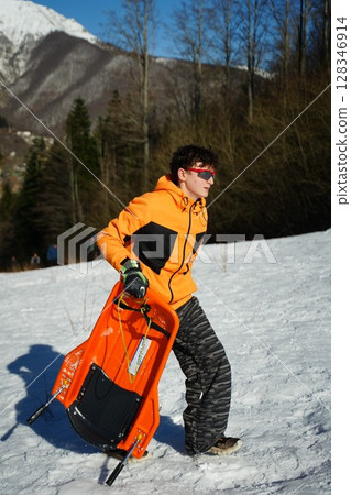 Teenager Carrying Orange Sled on Snowy Mountain Slope 128346914