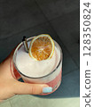 Hand holding pink summer cocktail with dried lemon slice and frothy foam in glass. Refreshing cold drink with stainless steel straw in sunlight. Trendy beverage for cafe or bar menu design. 128350824