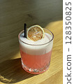 Refreshing pink summer cocktail in glass with frothy foam and dried lemon slice on wooden table. Bright sunlight and minimalist background. Vibrant mocktail or lemonade in trendy bar setting. 128350825