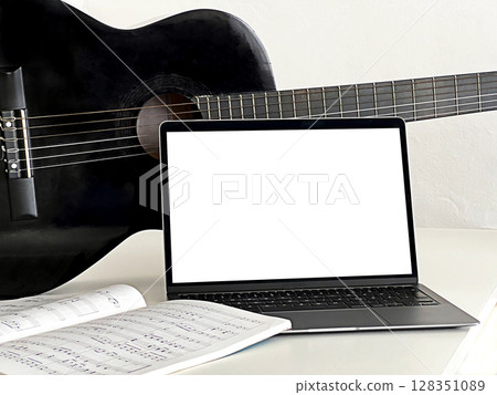 A black acoustic guitar rests beside a laptop displaying sheet music. A music book lies open on the table, A black acoustic guitar rests beside a laptop displaying sheet music. A music book lies open on the table, 128351089