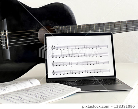 A black acoustic guitar rests beside a laptop displaying sheet music. A music book lies open on the table, A black acoustic guitar rests beside a laptop displaying sheet music. A music book lies open on the table, 128351090