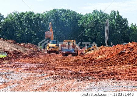Heavy machinery navigates muddy construction site surrounded by trees, showing ongoing development activity. Heavy machinery navigates muddy construction site surrounded by trees, showing ongoing development activity. 128351446