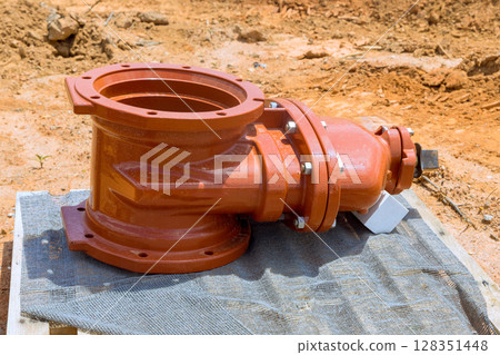 Large, orange pipe fire fitting rests waiting to be installed on wooden platform at construction site under works day 128351448