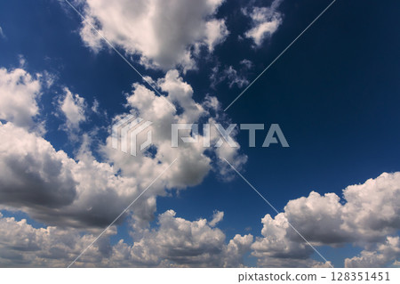 Fluffy white clouds float in deep blue sky during clear, sunny day, creating peaceful atmosphere. 128351451