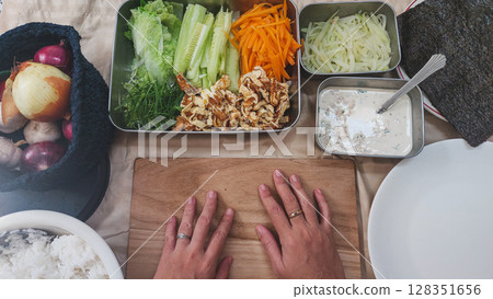 Hands positioned on a wooden board surrounded by fresh ingredients for making kimbap or gimbap which is a popular traditional korean food Hands positioned on a wooden board surrounded by fresh ingredients for making kimbap or gimbap which is a popular traditional korean food 128351656