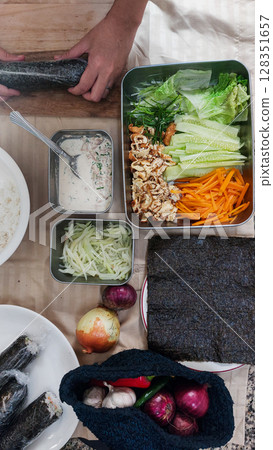 A vertical arrangement of ingredients while hands are rolling a gimbap or seaweed rice roll near vegetables and a sauce bowl A vertical arrangement of ingredients while hands are rolling a gimbap or seaweed rice roll near vegetables and a sauce bowl 128351657