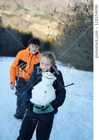 Teenager and Younger Sister Building Snowman Together 128354090