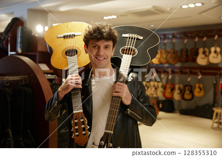 A man holds two guitars in front of a wall of various guitars 128355310