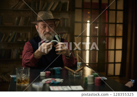 An elderly man engaged in a game of poker in a rustic setting, counting money 128355325