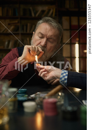 A man lights a cigarette, another man offers assistance, while playing poker 128355347