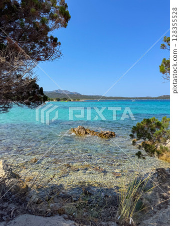 Idyllic beach scene featuring crystal-clear azure waters and a rocky shoreline framed by lush greenery. A perfect setting for relaxation and escape into nature's beauty.. Rena Bianca Sardinian  128355528