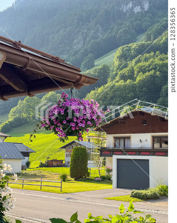 Vibrant floral basket hanging from a rooftop, set against a picturesque mountain village. The lush greenery and quaint houses provide a serene, idyllic backdrop on a sunny day. Austria  128356365