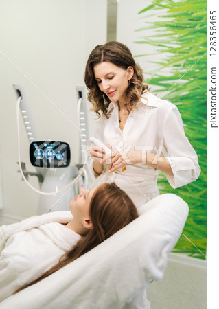Vertical shot of beautiful young woman receiving microneedling rejuvenation treatment in clinic of aesthetic medicine. Professional beautician performing needle mesotherapy on female client face. 128356465