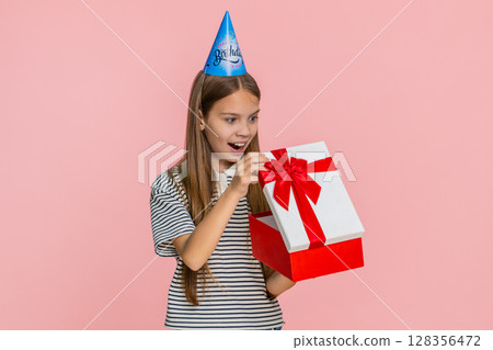 Child girl jumps with joy waves hands celebrating birthday while happily receiving surprise gift box 128356472