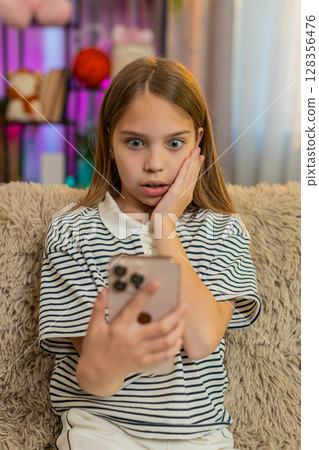 Child girl kid reads shocking message on smartphone with wide open eyes while sitting on home sofa 128356476