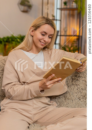 Adult woman smiling while reading interesting book at home enjoying relaxing peaceful moment on sofa Adult woman smiling while reading interesting book at home enjoying relaxing peaceful moment on sofa 128356480