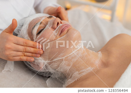 Woman completing facial wrap mask procedure in cosmetology clinic, beautician apply gauze skin care 128356494