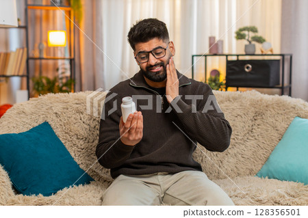Indian man at home holding cheek, suffering from toothache and searching for pain relief pills Indian man at home holding cheek, suffering from toothache and searching for pain relief pills 128356501