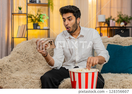 Indian man sitting on sofa, eating popcorn, watching video on smartphone focused enjoying relaxation 128356607