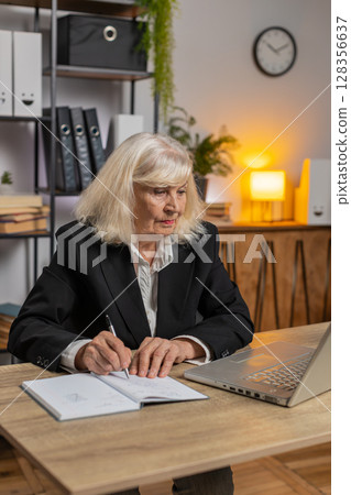 Senior Caucasian businesswoman using laptop computer and writing notes in diary at home office desk Senior Caucasian businesswoman using laptop computer and writing notes in diary at home office desk 128356637