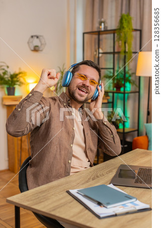 Happy Caucasian man in headphones and sunglasses dancing listening to music at home office desk 128356685