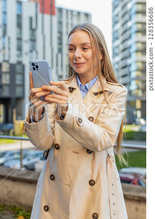 Smiling mature businesswoman chatting on internet via mobile phone standing on downtown city street 128356686