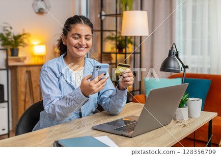 Happy Indian woman freelancer using credit card and smartphone for online shopping at table 128356709