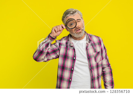 Mature man holds magnifying glass near face looking at camera with big zoomed eye, analyzing Mature man holds magnifying glass near face looking at camera with big zoomed eye, analyzing 128356710