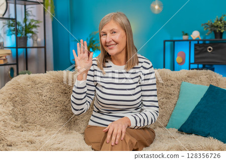 Mature woman waving hello hi on sofa, smiling warmly, expressing friendliness and openness at home Mature woman waving hello hi on sofa, smiling warmly, expressing friendliness and openness at home 128356726