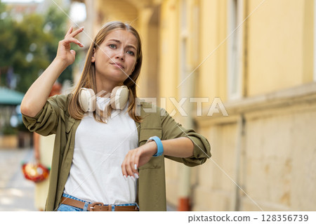 Serious dissatisfied young Caucasian woman standing and looking for someone waiting on city street Serious dissatisfied young Caucasian woman standing and looking for someone waiting on city street 128356739
