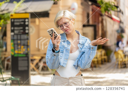 Upset young woman using cellphone typing browsing, loses becoming shocked bad news on city street Upset young woman using cellphone typing browsing, loses becoming shocked bad news on city street 128356741