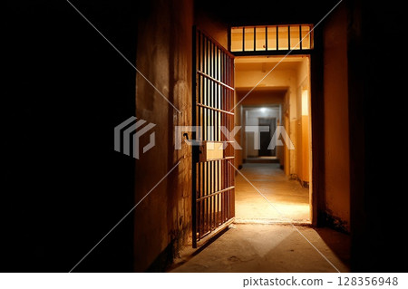 Open prison cell door revealing dimly lit corridor with shadows and textured walls creating atmosphere Open prison cell door revealing dimly lit corridor with shadows and textured walls creating atmosphere 128356948
