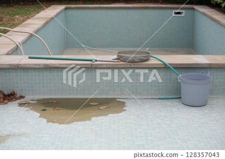Empty swimming pool with cleaning equipment and water accumulation on tiled surface 128357043
