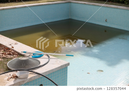 Unmaintained swimming pool with green water and cleaning equipment on the edge of the pool 128357044