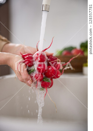 Close up view of a woman washes a bunch of red radishes under the pressure of flowing water in the white kitchen, a basket with tomato, broccoli and onions in the background, grapes Close up view of a woman washes a bunch of red radishes under the pressure of flowing water in the white kitchen, a basket with tomato, broccoli and onions in the background, grapes 128357781