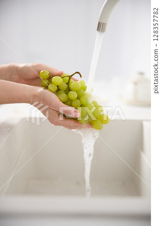 Close up view of a woman washes fresh green grapes under the pressure of flowing water in the white kitchen Close up view of a woman washes fresh green grapes under the pressure of flowing water in the white kitchen 128357782
