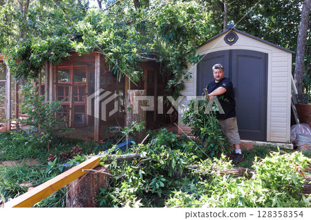 Person clears branches leaves around shed while maintaining the garden in afternoon. 128358354