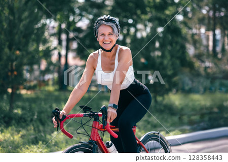 Happy sporty woman in helmet and white top cycling in park and smiling at camera 128358443