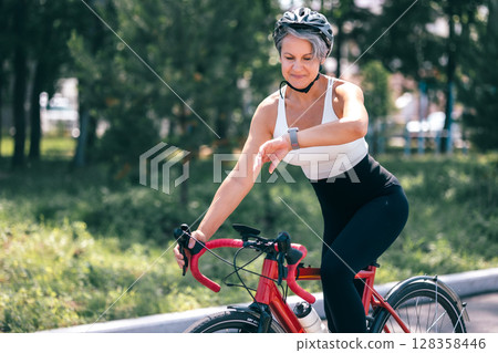 Senior Woman Riding Bicycle and Checking Smartwatch During Outdoor Workout Senior Woman Riding Bicycle and Checking Smartwatch During Outdoor Workout 128358446