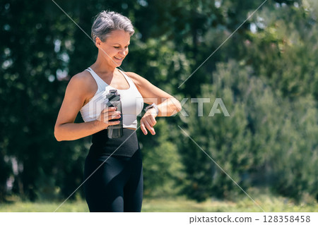Smiling Mature Woman Checking Fitness Watch While Holding Water Bottle After Outdoor Workout 128358458