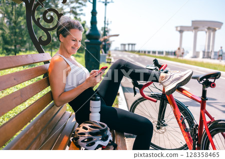 Senior Woman Relaxing on Bench with Smartphone After Bike Ride on Sunny Promenade 128358465