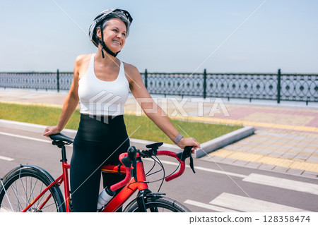 Smiling Senior Woman Standing with Bicycle on Seafront Road on Bright Sunny Day 128358474