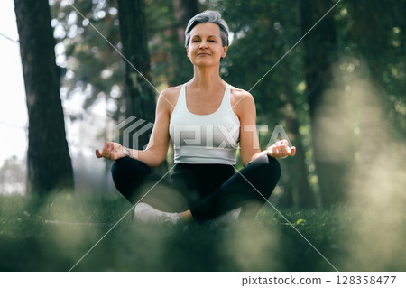 Serene Mature Woman in Sukhasana Yoga Pose Meditating Peacefully in Forest Nature Setting Serene Mature Woman in Sukhasana Yoga Pose Meditating Peacefully in Forest Nature Setting 128358477