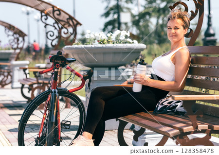 Fit woman resting on bench with bicycle and water bottle during summer ride in urban park Fit woman resting on bench with bicycle and water bottle during summer ride in urban park 128358478