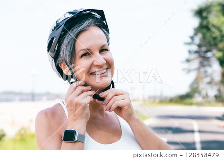 Active smiling woman adjusting bike helmet on a bright sunny day before outdoor exercise ride 128358479